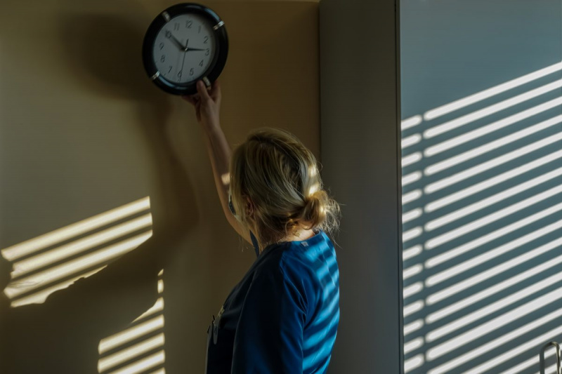 The windows in the patient room of a hospital are closed with blinds. A nurse holds a wall clock up with one arm to hang it on the wall. Her body with the clock casts a shadow to the left. Light shows on a wardrobe to the right and left over her shadow. There are also elongated ones on her back that look like the scan of an AI. Photo © Michael Nguyen/VG Bild-Kunst, Bonn